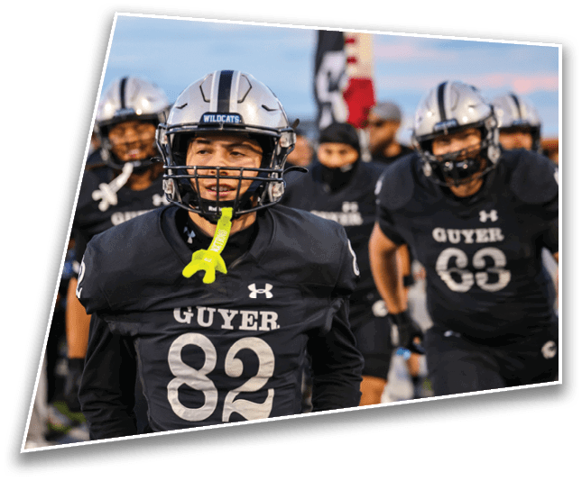 People wearing football uniforms with helmets and face paint are standing together on a field, with one person in the foreground smiling and holding a mouthguard. Description generated by AI