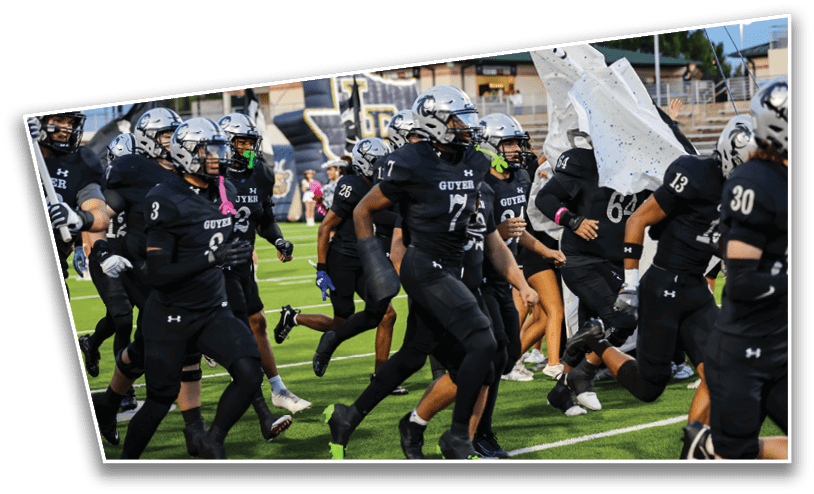 A group of people wearing black football uniforms and helmets are running onto a field through torn white paper banners. Description generated by AI