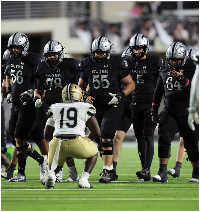 People wearing black football uniforms with silver helmets are on a field, with one player in the center wearing number 55. Description generated by AI