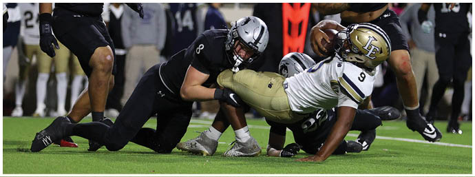 Two football players in black uniforms with silver helmets are on a field with spectators in the background, one player wearing number 99 and the other wearing number 9. Description generated by AI