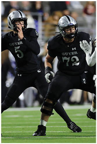 Two people in black football uniforms with helmets are positioned on a field, one preparing to throw a ball and the other in a blocking stance. Description generated by AI