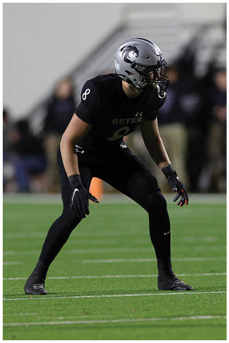A person in black athletic gear and a silver helmet is poised in a ready stance on a football field during a game. Description generated by AI
