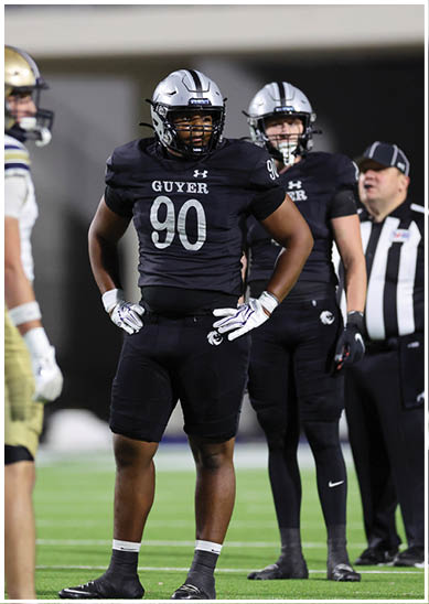 Two people wearing black football uniforms with the word “GUYER" and the number 90 on the front stand on a field with their hands on their hips. Description generated by AI