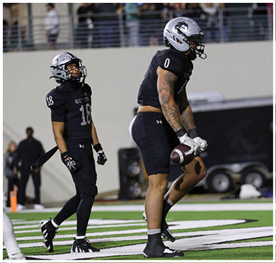 An adult wearing a black football uniform and silver helmet is holding a football on a field with a trailer and spectators in the background. Description generated by AI