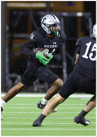A person wearing a black football uniform with the word “GUYER" and a silver helmet is holding a football and appears to be running on a field at night. Description generated by AI
