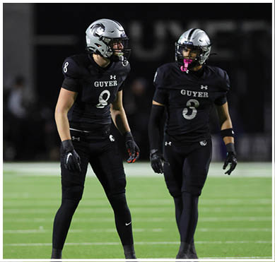 Two people in black football uniforms stand on a green field with white yard lines, one wearing number 8 and the other partially visible wearing number 3. Description generated by AI