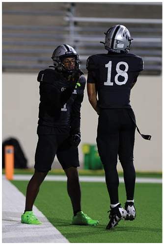 A person wearing a black football uniform and helmet is standing on a field, appearing to gesture with one hand near their face. Description generated by AI