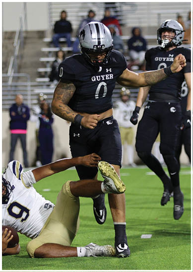 A person wearing a black football uniform with the number 0 and a silver helmet is running on a field while another person reaches to grab their leg. Description generated by AI