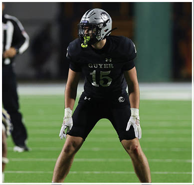A person wearing a black football uniform with the number 15 and the word “GUYER" stands on a green field with knees slightly bent and hands in white gloves positioned in front. Description generated by AI