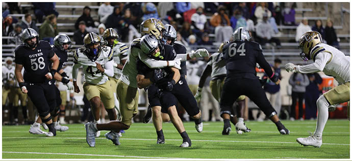 People in football uniforms are actively playing on a field with one player tackling another while others run and watch, with spectators seated in the background. Description generated by AI