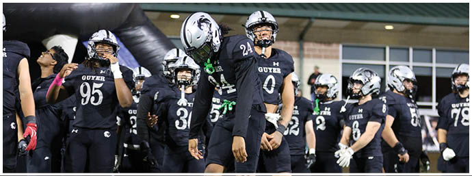A group of people wearing black football uniforms and helmets stand together on a field near a building. Description generated by AI