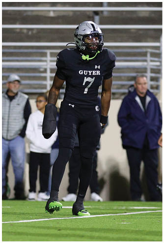A person wearing a black football uniform with the number 7 and the word “GUYER" on the front is standing on a football field with another player and people in the background. Description generated by AI