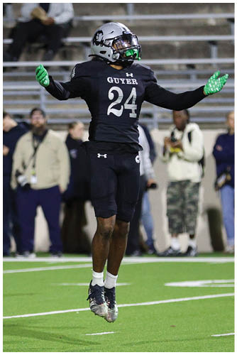 A person wearing a black football uniform with the number 24 and green gloves is jumping on a football field with spectators in the background. Description generated by AI