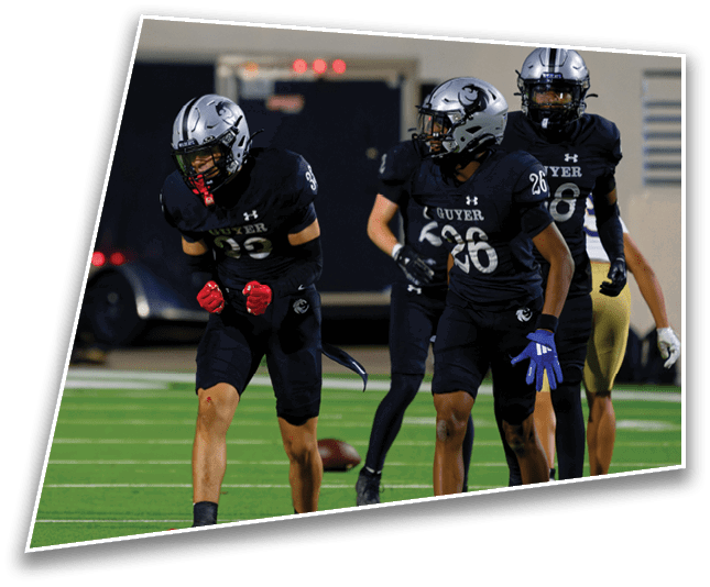 Three people in black football uniforms and helmets are on a green field with one person clenching fists and the others walking nearby. Description generated by AI