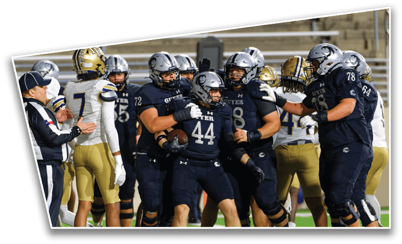 A group of football players in dark uniforms are celebrating on the field while players in white and gold uniforms stand nearby. Description generated by AI