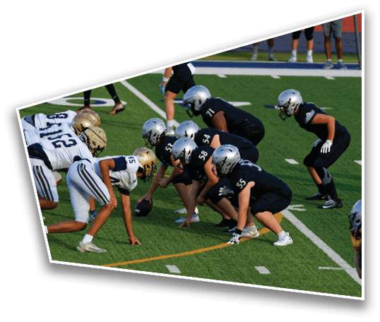 Two opposing American football teams in helmets and uniforms are lined up facing each other on a green field, preparing for a play. Description generated by AI