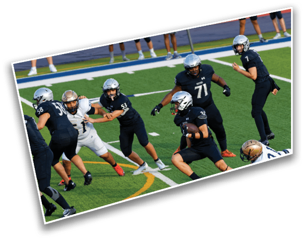 A group of people wearing helmets and sports uniforms are playing football on a green field with one person running while holding the ball and others preparing to block or tackle. Description generated by AI