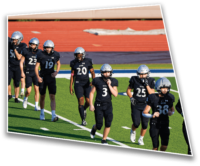 Five people wearing black football uniforms and silver helmets are walking on a green field near a running track. Description generated by AI