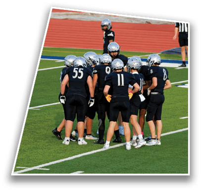 A group of people wearing black sports uniforms and helmets are huddled together on a green football field with a red running track in the background. Description generated by AI