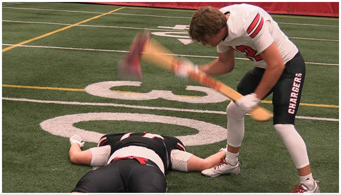 One person lies face down on an indoor sports field while another person stands over them holding a blurred wooden bat in a raised position. Description generated by AI