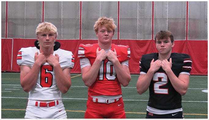 Three adults wearing different colored football uniforms stand side by side on an indoor field with their hands clasped near their chests. Description generated by AI