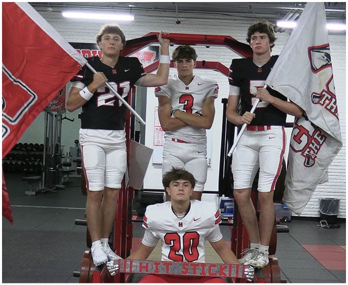 Four people in football uniforms pose in a gym with two holding flags and one sitting on a bench holding a sign that says “HIT STICK!!!" Description generated by AI