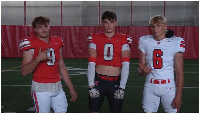 Three people wearing football uniforms stand on an indoor field with two in red jerseys and one in a white jersey, each making hand gestures. Description generated by AI
