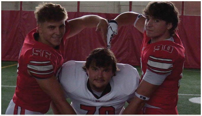 Three people in football uniforms pose together on an indoor field with two wearing red jerseys and one in a white jersey. Description generated by AI
