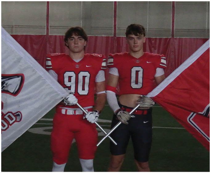 Two adults wearing red football jerseys hold flags while standing indoors on a sports field. Description generated by AI