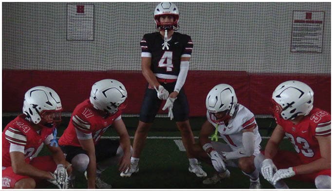 Five people in football uniforms and helmets are positioned on a field with one standing in the center and four crouching around them. Description generated by AI