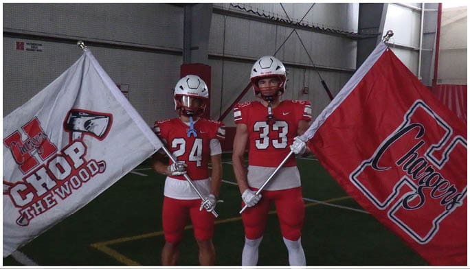 Two people in red football uniforms and helmets stand indoors holding large flags with team logos and slogans. Description generated by AI