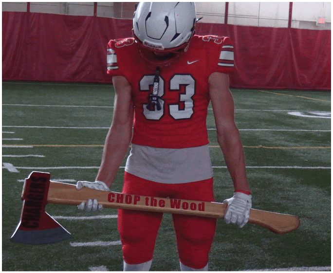 A person wearing a red football uniform and white helmet is holding a large wooden axe with the words “CHOP the Wood" written on the handle while standing on an indoor football field. Description generated by AI