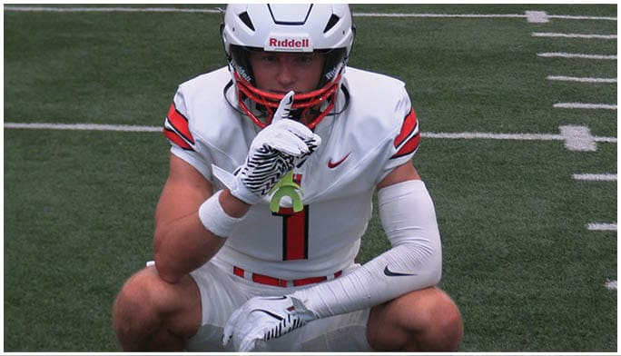 A person in a white football uniform with red and black accents is crouching on a field while holding a finger to their lips in a shushing gesture. Description generated by AI