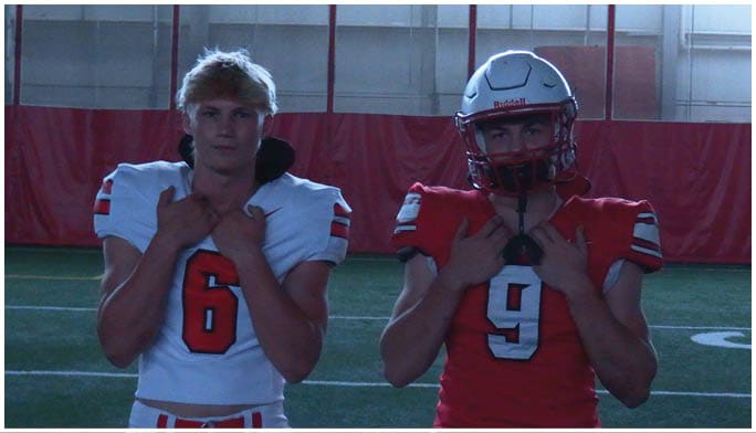 Two people wearing football uniforms stand side by side indoors on a field, one in a white jersey with the number 6 and the other in a red jersey with the number 9 and a helmet. Description generated by AI
