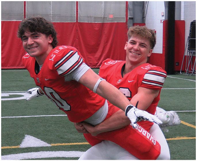 Two people wearing red football jerseys are smiling and playfully posing on an indoor football field, with one person lifting the other. Description generated by AI
