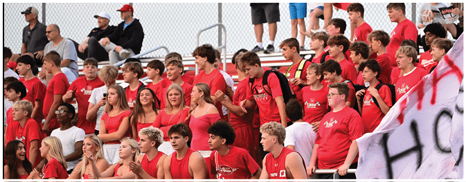 A person wearing red shorts and a dark shirt is in the foreground while a child in a white shirt stands near a chain-link fence in the background. Description generated by AI