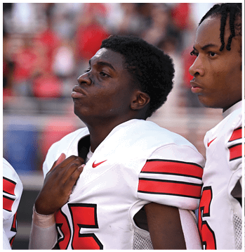 A person wearing a white sports jersey with red and black stripes on the sleeve and a red number is holding the collar near their chest. Description generated by AI