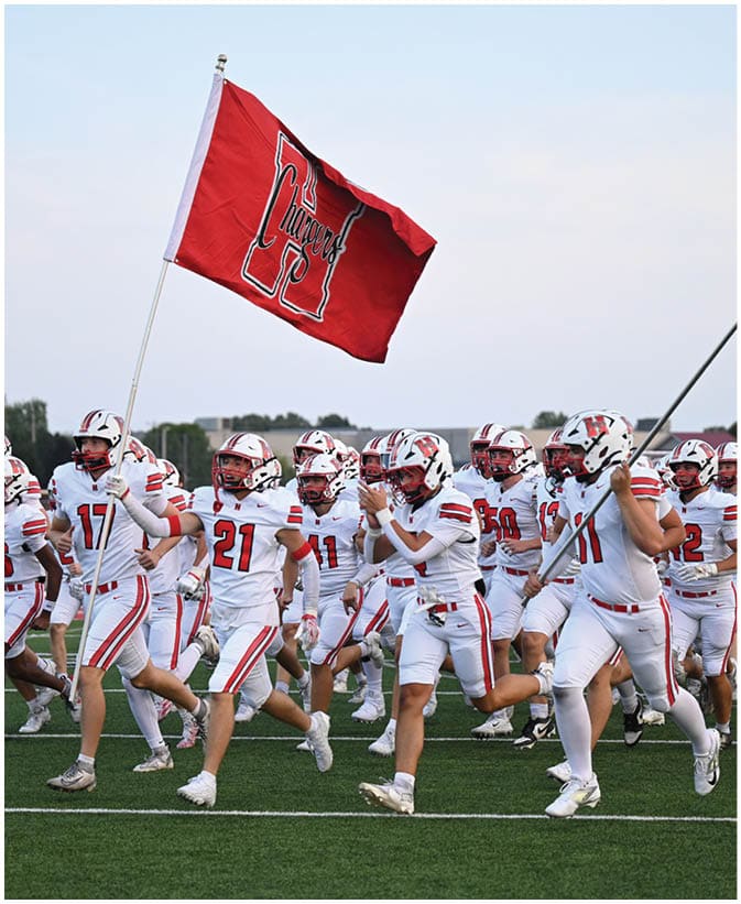 A group of people in white sports uniforms with helmets are running on a field, with one person holding a red flag that has a large letter and text on it. Description generated by AI