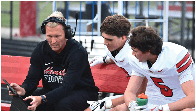 An adult wearing a headset and black sportswear is showing a tablet to two people in white football uniforms sitting on a bench on a sports field. Description generated by AI