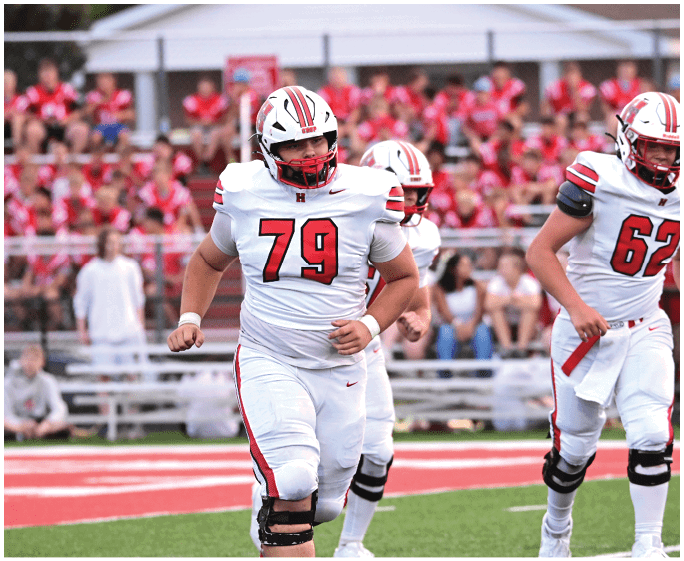 Three people in white football uniforms with red and black accents are running on a football field with a crowd in the background. Description generated by AI