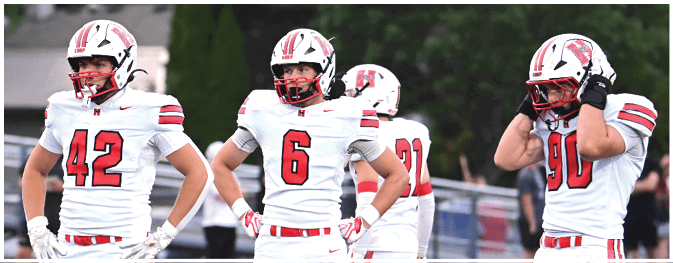 Three people in white football uniforms with red and black accents stand on a field with trees and spectators in the background. Description generated by AI