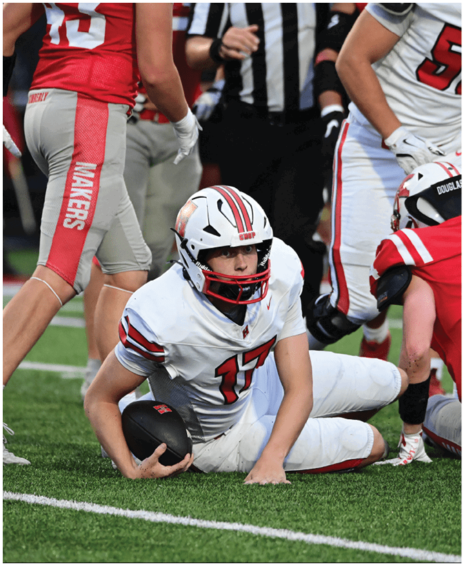 A person wearing a white football uniform with the number 17 is lying on the ground holding a football while surrounded by other players in red and white uniforms during a game. Description generated by AI