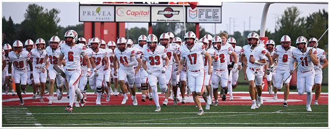 A group of people wearing white football uniforms with red accents are running together on a field with a scoreboard and goalposts visible in the background. Description generated by AI