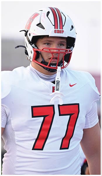 A person wearing a white football uniform with the number 77 in red stands in the foreground while another person in a white and red football uniform is visible in the background. Description generated by AI