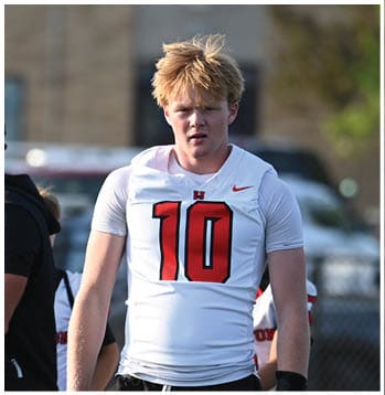 A person wearing a white sports jersey with the number 10 and black shorts is walking on a sports field with another person in black clothing holding papers nearby. Description generated by AI