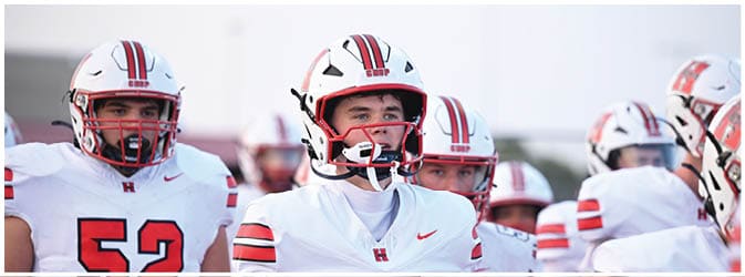 People wearing white football uniforms and helmets are standing together on a field. Description generated by AI