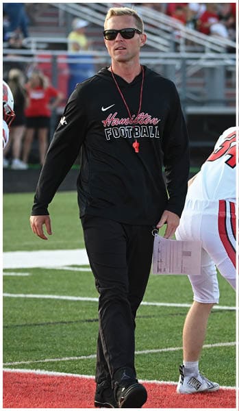 An adult wearing sunglasses and a black Hamilton Football outfit walks on a football field while holding a clipboard near a person in white football gear bending forward. Description generated by AI
