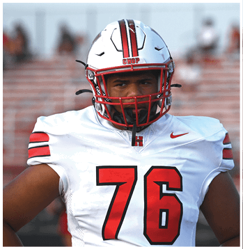 A person wearing a white football uniform with red and black stripes and the number 76 on the front is standing on a field with blurred bleachers in the background. Description generated by AI