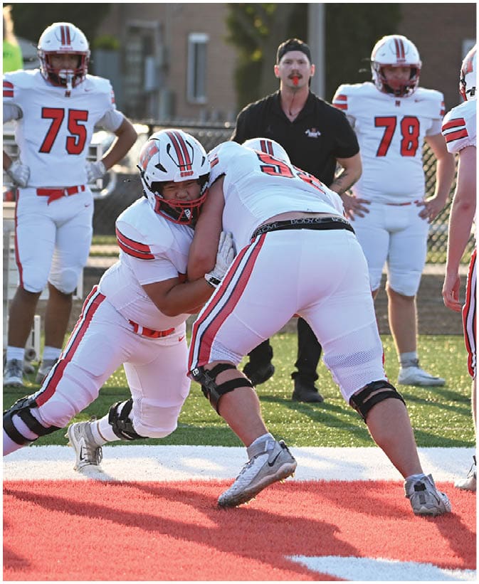 Two people in football uniforms are engaged in a blocking drill on a field while others in similar uniforms and a coach with a whistle observe in the background. Description generated by AI