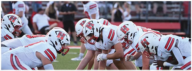 People in football uniforms and helmets are lined up on a field in a ready stance, preparing for a play. Description generated by AI